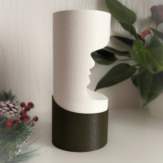 Decorative vase with textured white top and black base on a surface with plants and pinecones in the background.